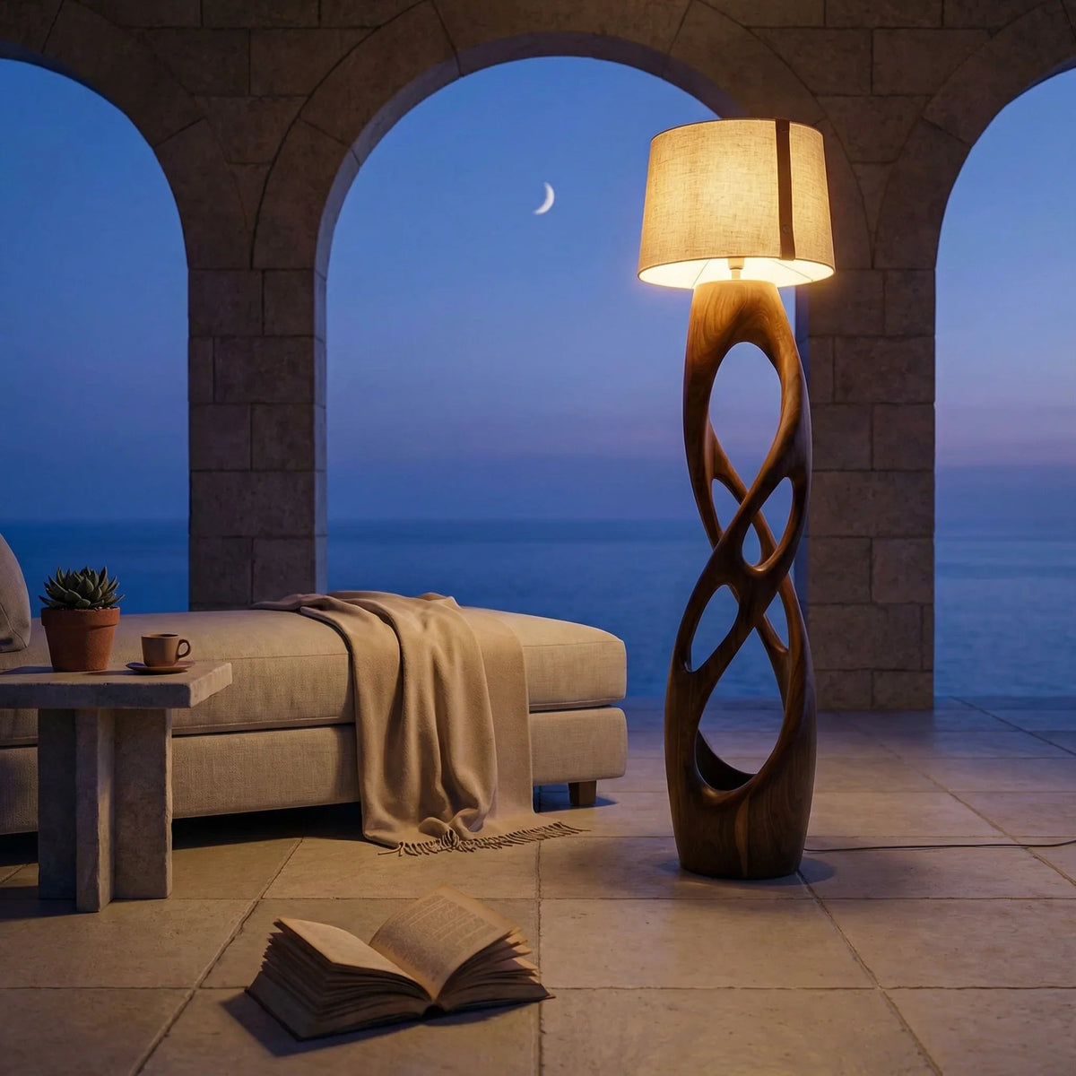 Terrasse einer Mittelmeer-Villa in der Dämmerung mit skulptierter Holz-Stehlampe, Loungesessel, aufgeschlagenem Buch und Meerblick.